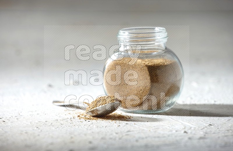 A glass spice jar and metal spoon full of cumin powder on textured white flooring