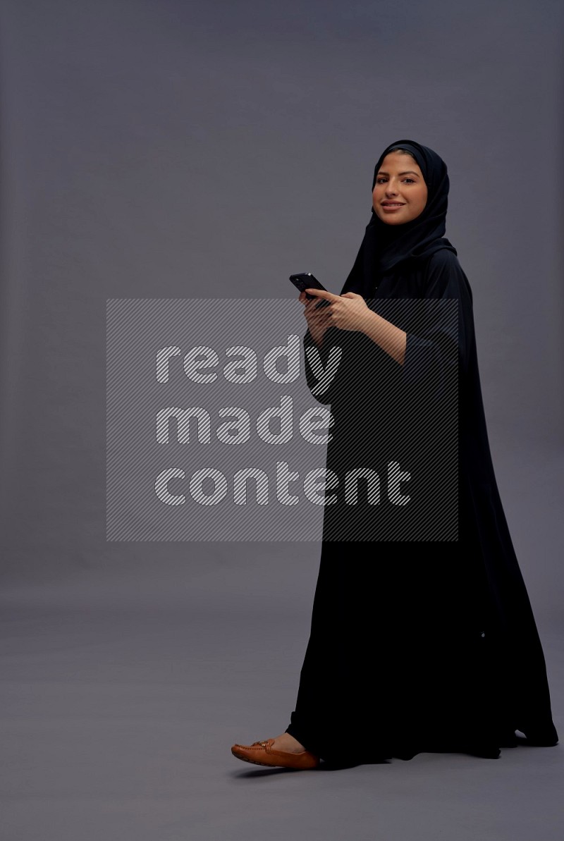 Saudi woman wearing Abaya standing texting on phone on gray background