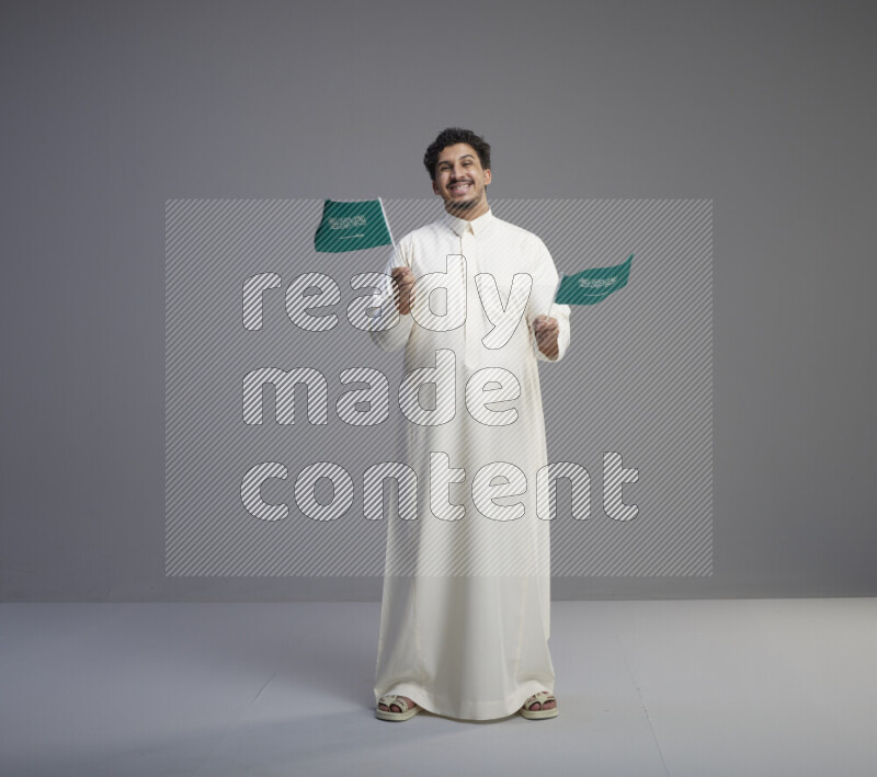 A Saudi man standing wearing thob holding small Saudi flag on gray background