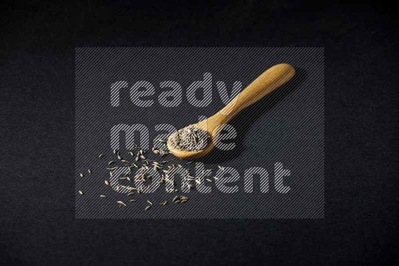 A wooden spoon full of cumin seeds on black flooring