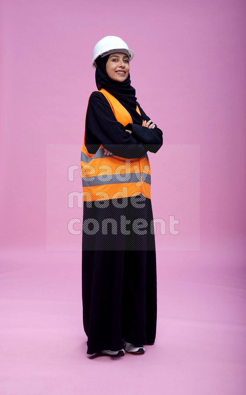 Saudi woman wearing Abaya with engineer vest and helmet standing with crossed arms on pink background