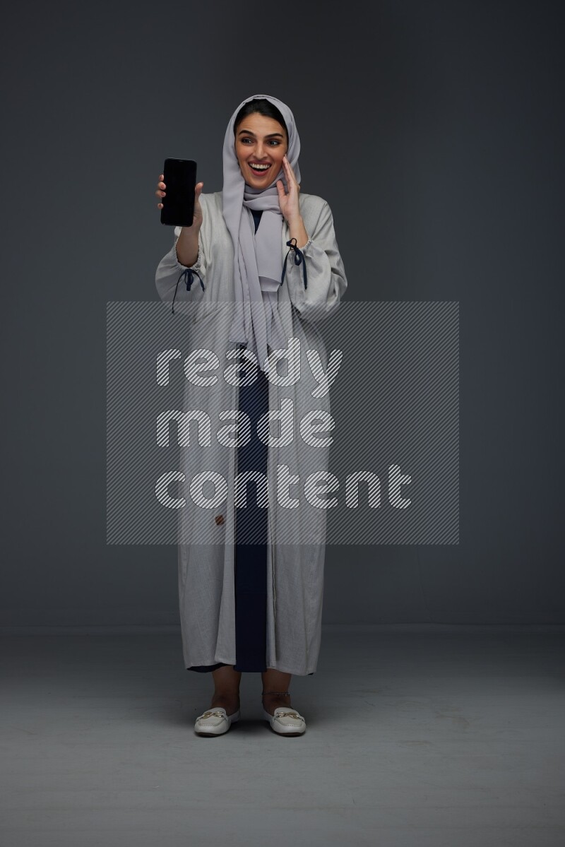 A Saudi woman wearing a light gray Abaya and head scarf standing and showing the phone's screen while holding a blue luggage eye level on a grey background