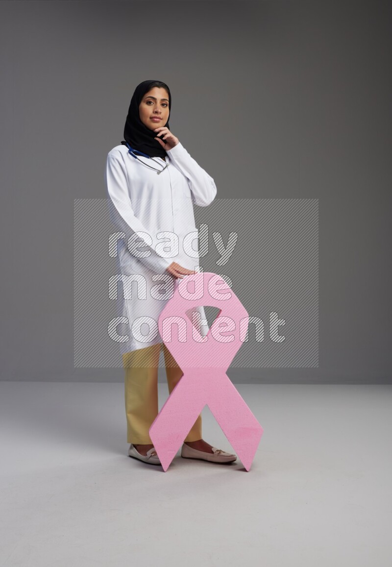 Saudi woman wearing lab coat with stethoscope standing holding awareness ribbon symbol standing on Gray background