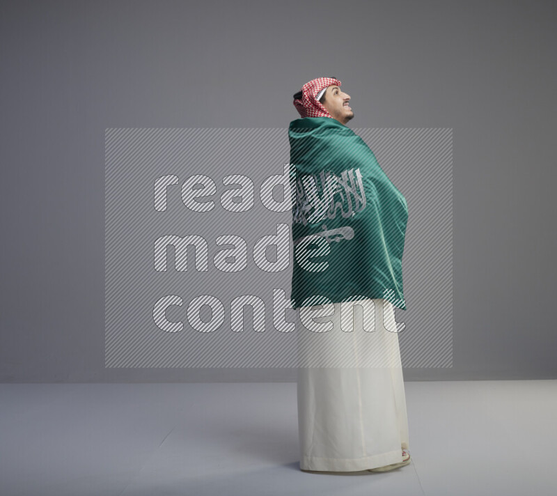 A Saudi man standing wearing thob and red shomag wrapping big saudi flag on gray background