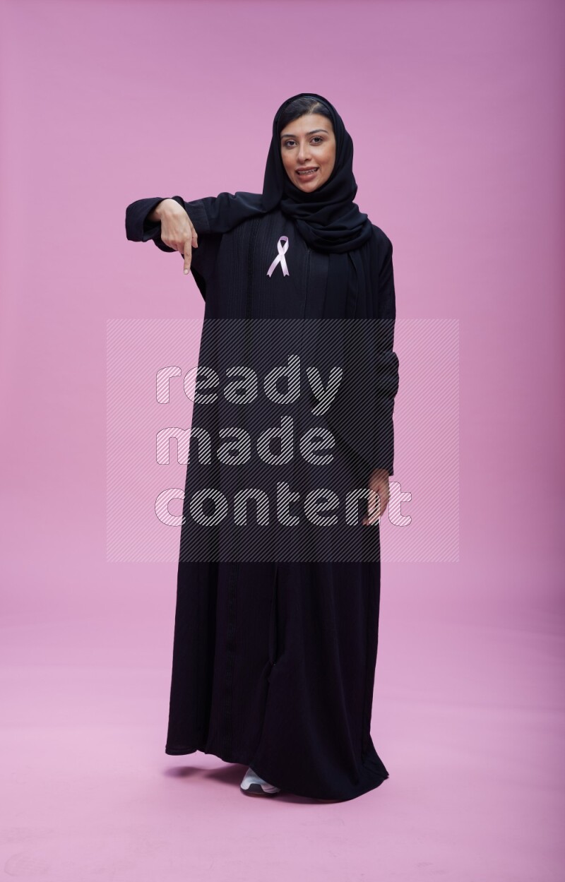 Saudi woman wearing pink ribbon on Abaya standing interacting with the camera on pink background