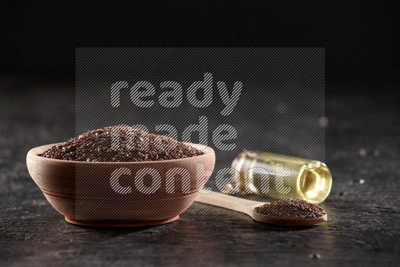 A wooden bowl and spoon full of flax and a glass jar of flax oil on a textured black flooring in different angles