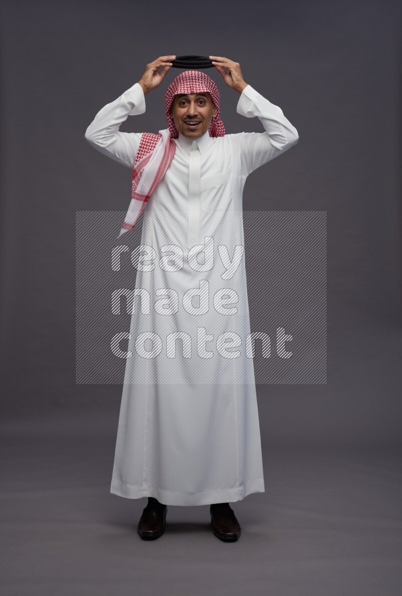 Saudi man wearing thob and shomag standing hands behind head on gray background