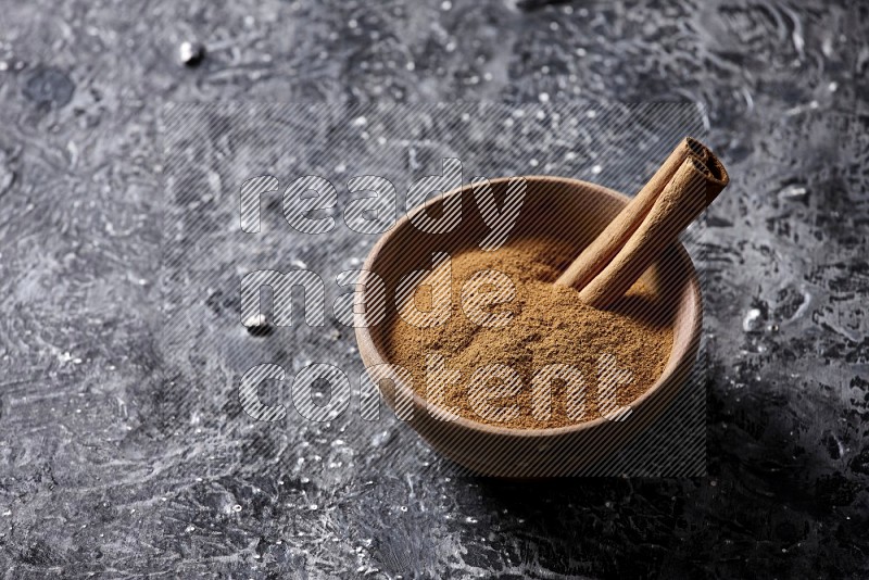 Wooden bowl full of cinnamon powder and a cinnamon stick on a textured black background