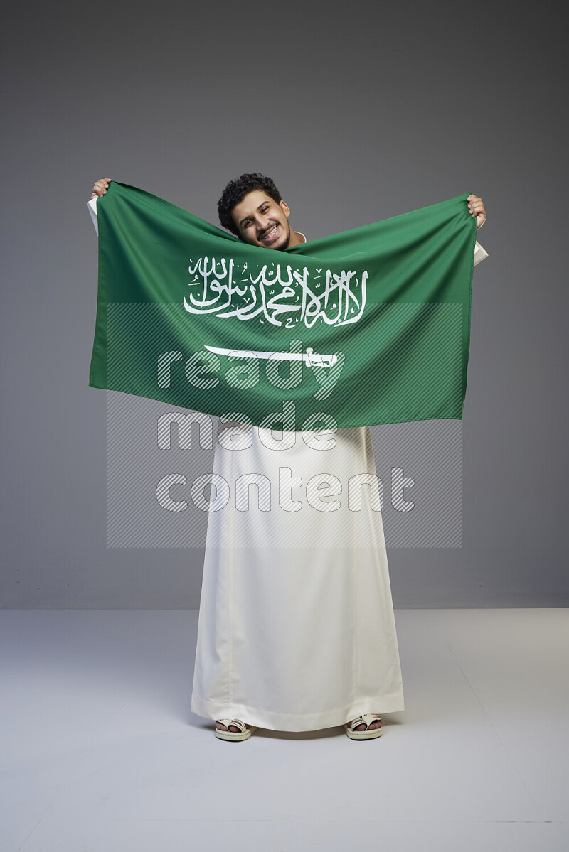 A Saudi man standing wearing thob holding big Saudi flag on gray background