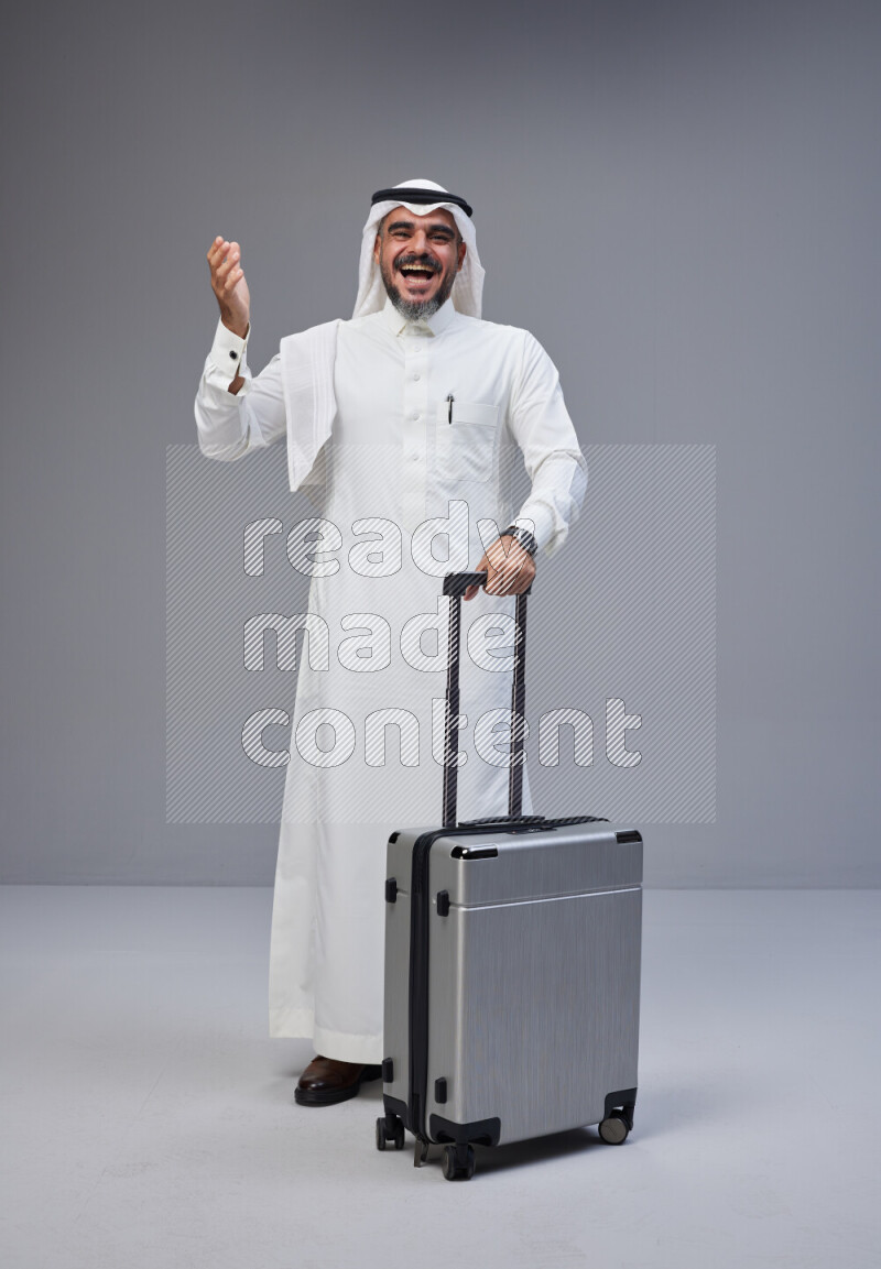 Saudi man wearing Thob and white Shomag standing holding Travel bag on Gray background