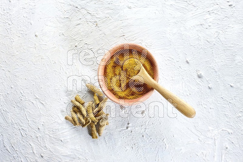 A wooden bowl and wooden spoon full of turmeric powder with dried turmeric fingers beside it on textured white flooring