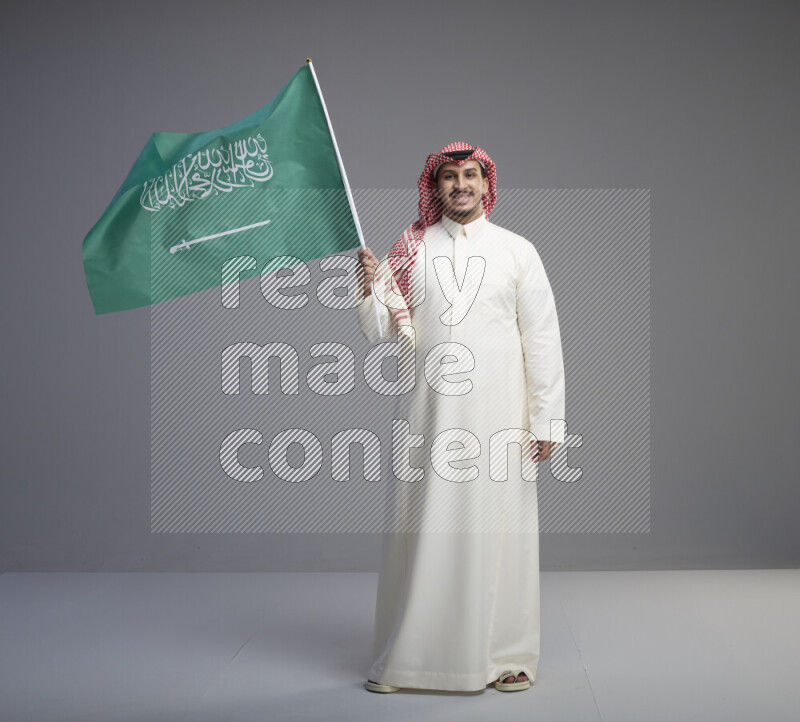 A Saudi man standing wearing thob and red shomag raising big Saudi flag on gray background