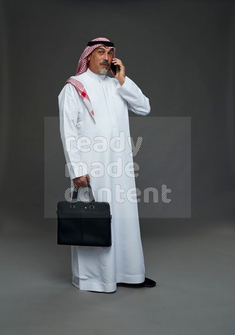 Saudi man with shomag Standing holding bag talking on phone on gray background