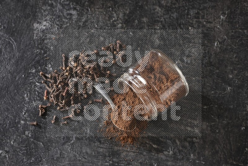 A flipped glass jar and metal spoon full of cloves powder with cloves spread on a textured black flooring