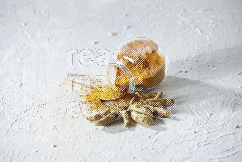 A flipped glass spice jar and metal spoon full of turmeric powder and powder spilled out of it with dried whole fingers on textured white flooring