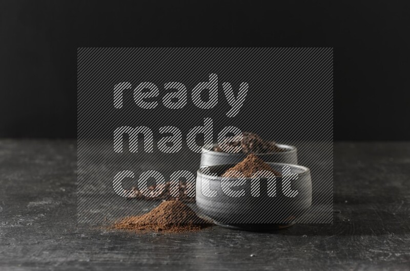 2 Black pottery bowls full of cloves and the other full of cloves powder on textured black flooring