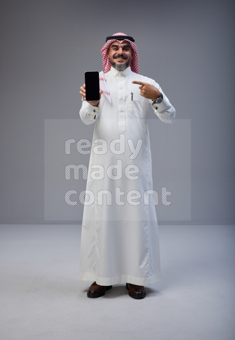 Saudi man Wearing Thob and red Shomag standing showing phone to camera on Gray background