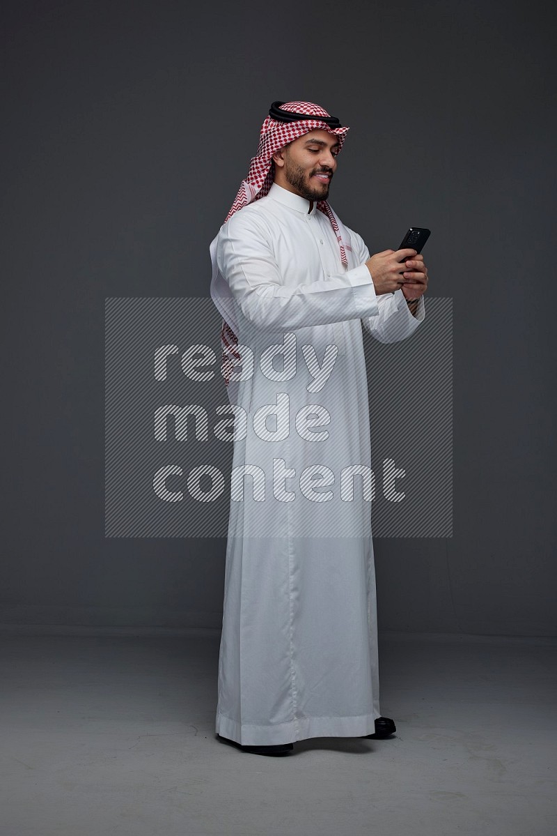 A Saudi man wearing Thobe and Shmagh standing and using his phone eye level on a gray background