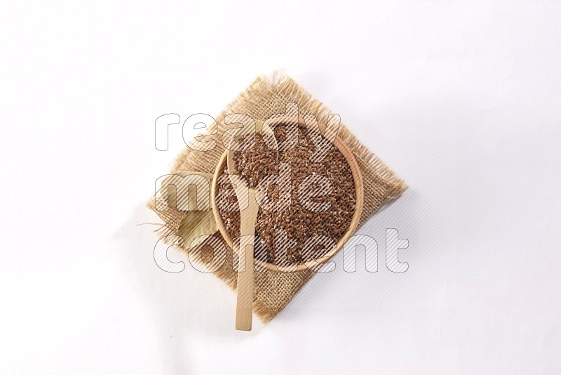 A wooden bowl full of flax seeds and a wooden spoon full of the seeds on burlap fabric on a white flooring