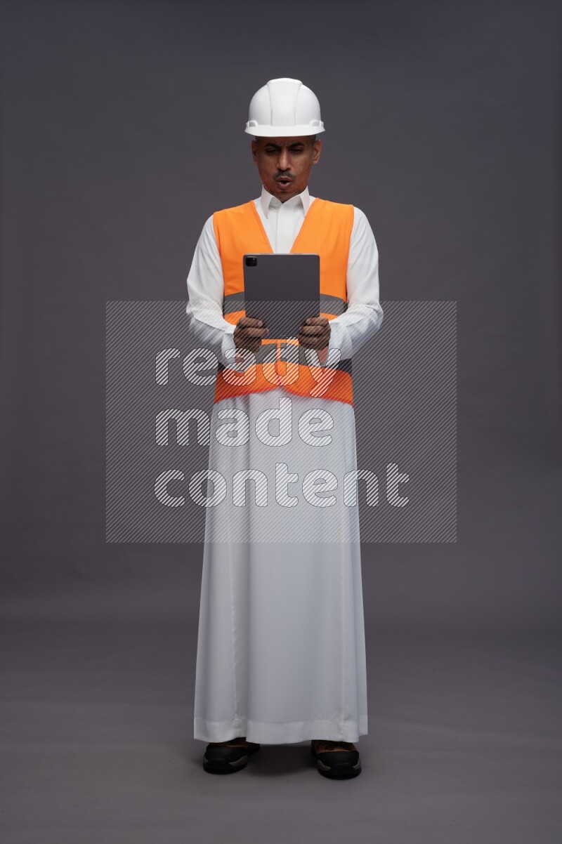 Saudi man wearing thob with engineer vest standing working on tablet on gray background