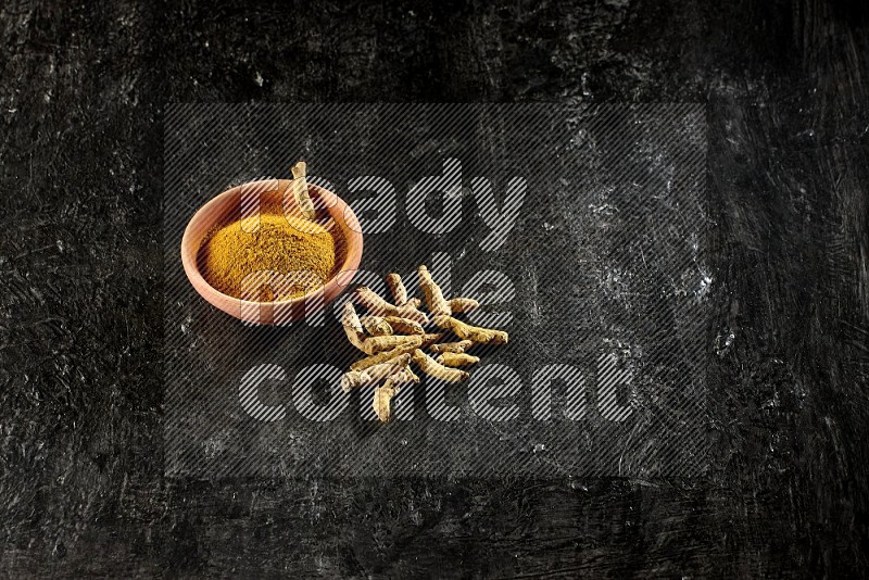 A wooden bowl full of turmeric powder with dried turmeric whole fingers on textured black flooring
