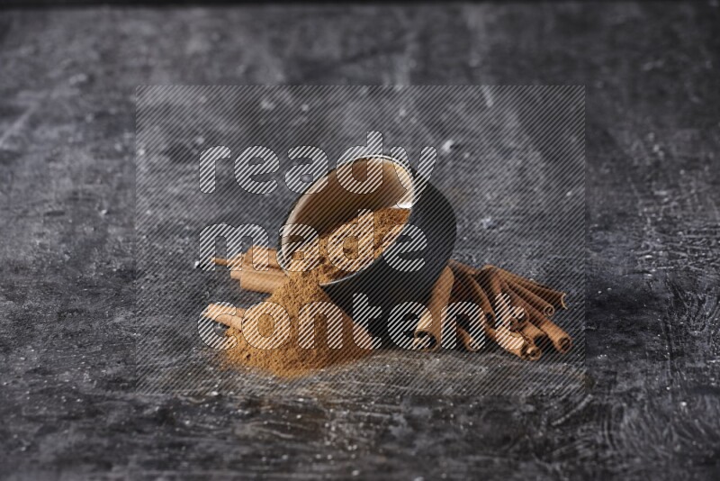 Black pottery bowl over filled with cinnamon powder and cinnamon sticks around the bowl on a textured black background