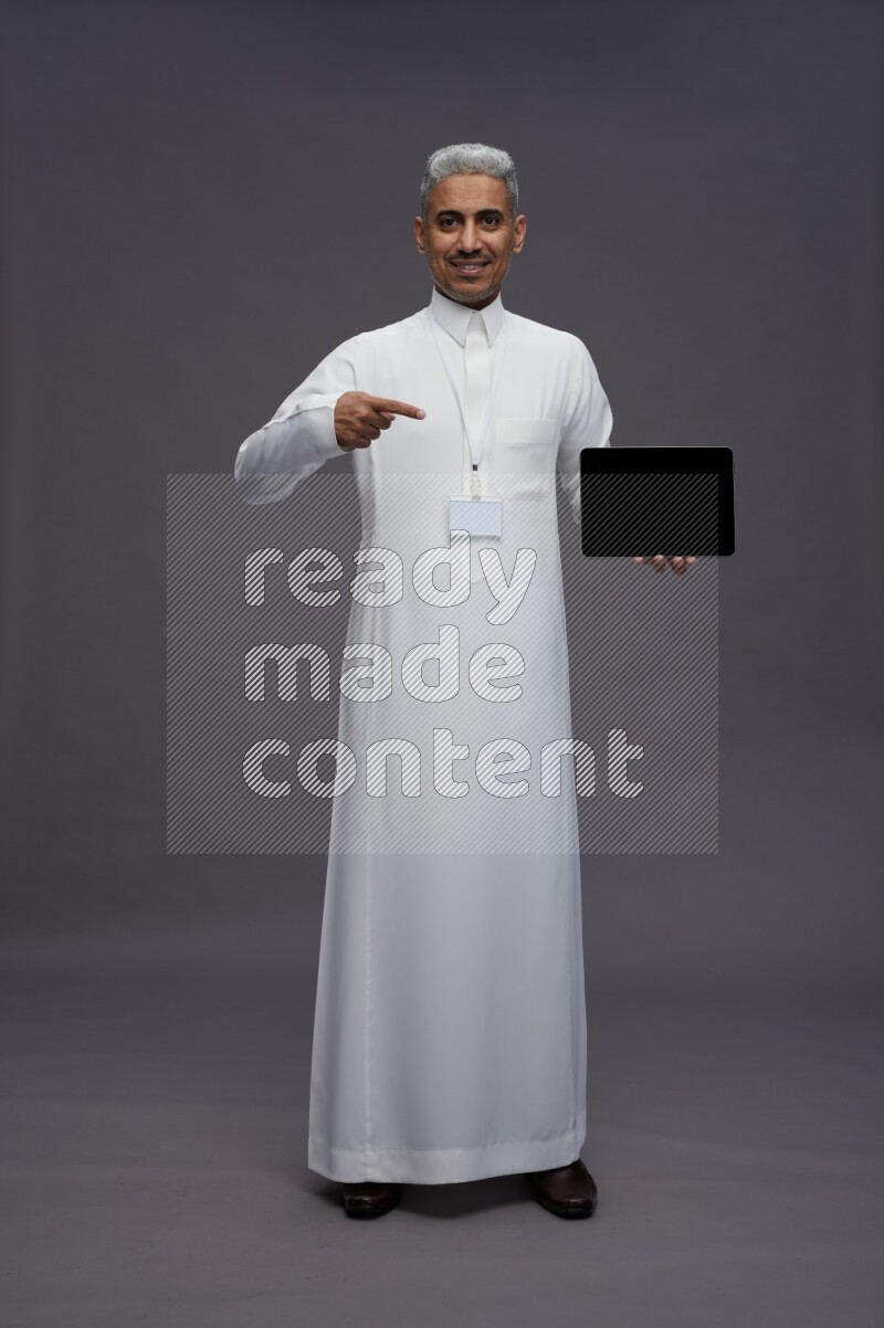Saudi man wearing thob with neck strap employee badge standing showing tablet to camera on gray background
