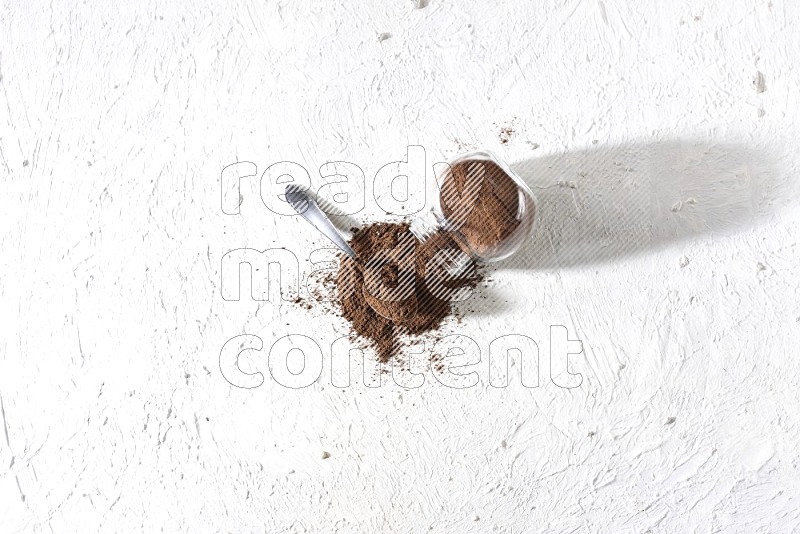 A flipped glass spice jar and a metal spoon full of cloves powder and powder came out of the jar on textured white flooring