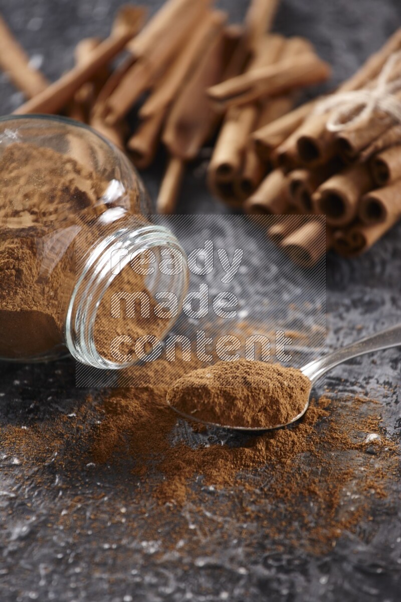 Herbal glass jar full cinnamon powder flipped and a metal spoon full of powder, cinnamon sticks stacked and bounded in the back on textured black background in different angles