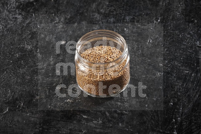 A glass jar full of mustard seeds on a textured black flooring
