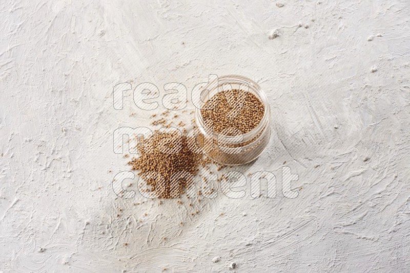 A glass jar full of mustard seeds and more seeds spread on a textured white flooring