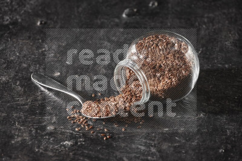 A glass spice jar full of flaxseeds flipped and seeds spread out with a metal spoon full of the seeds on a textured black flooring