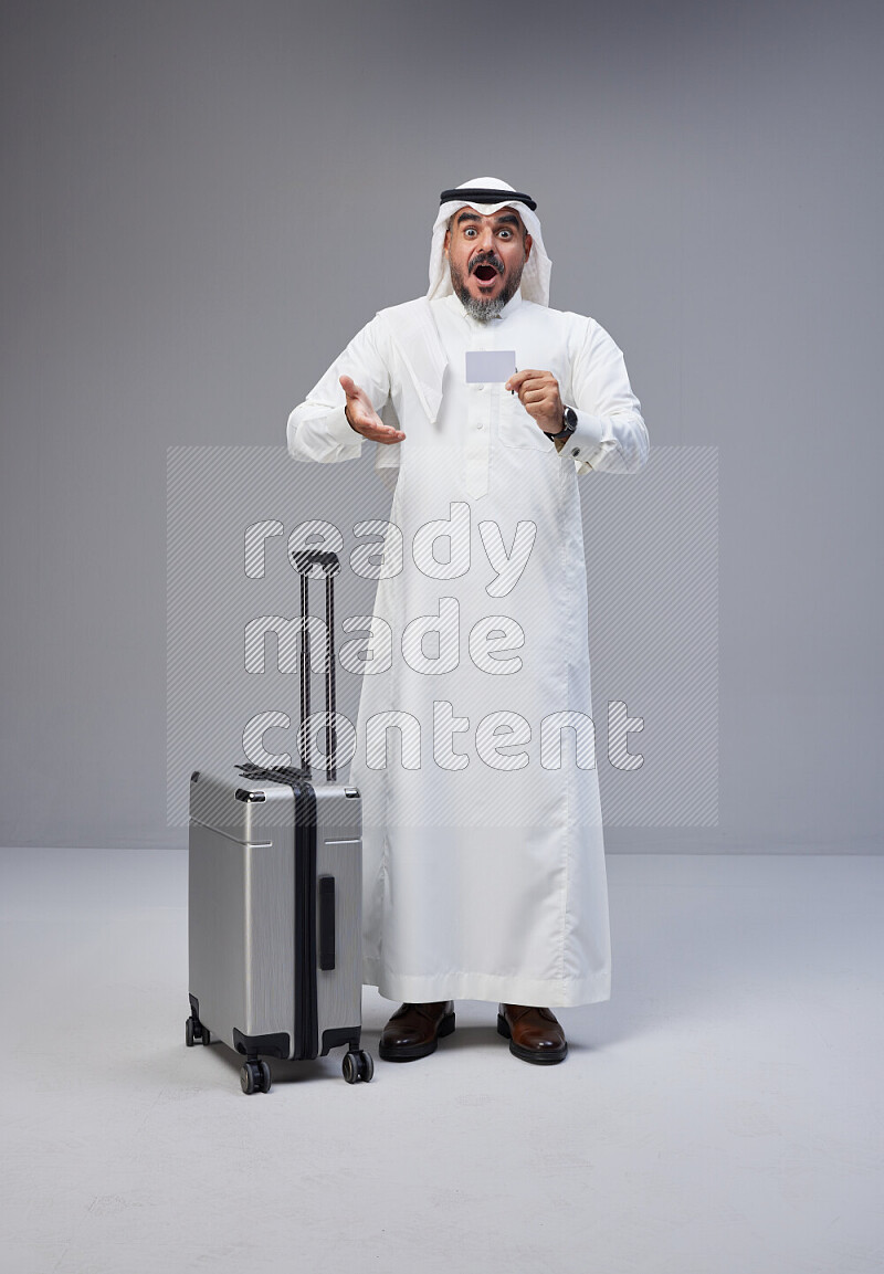 Saudi man wearing Thob and white Shomag standing holding Travel bag and ATM card on Gray background