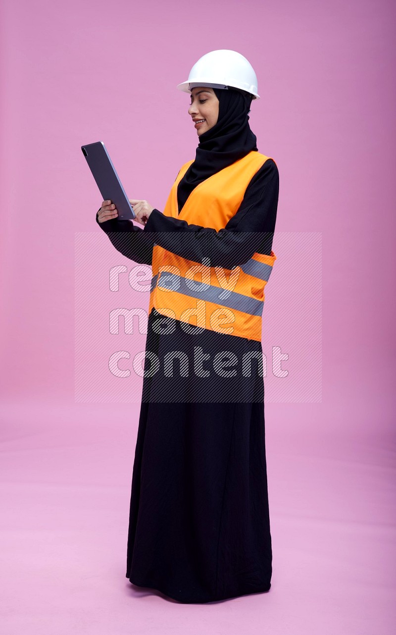 Saudi woman wearing Abaya with engineer vest and helmet standing working on tablet on pink background