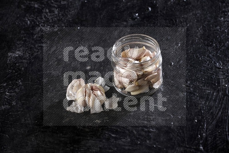 A glass jar full of garlic cloves and beside it a garlic bulb on a textured black flooring in different angles