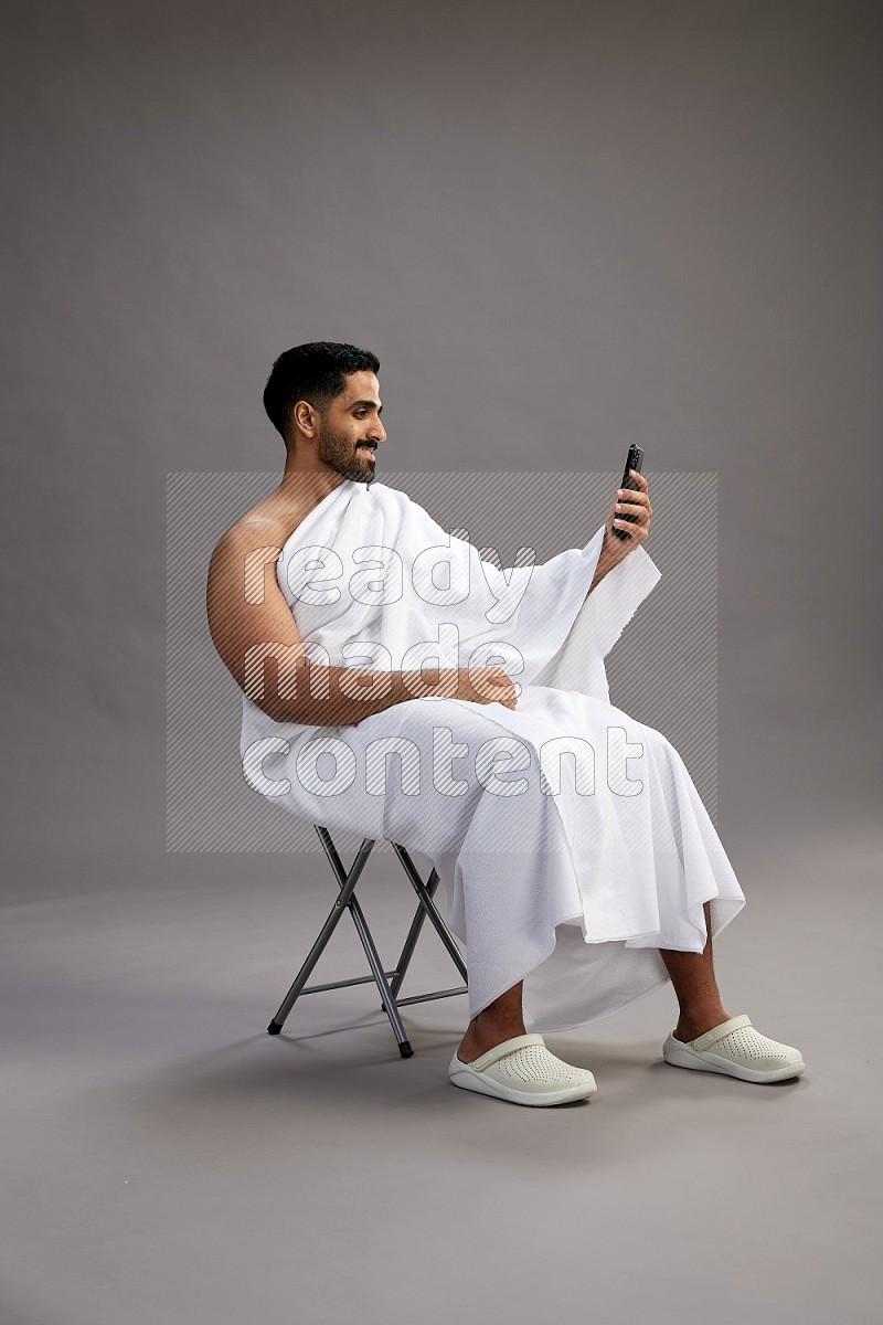 A man wearing Ehram sitting on chair taking selfie on gray background
