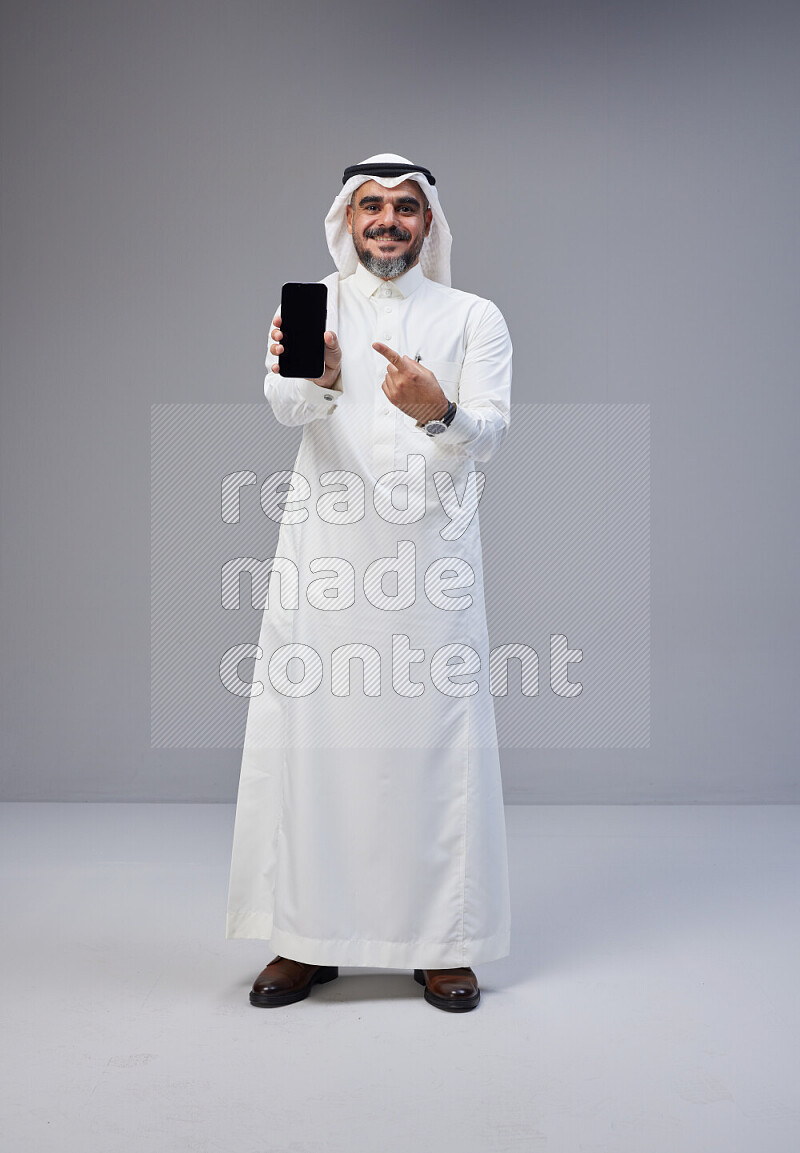 Saudi man Wearing Thob and white Shomag standing showing phone to camera on Gray background