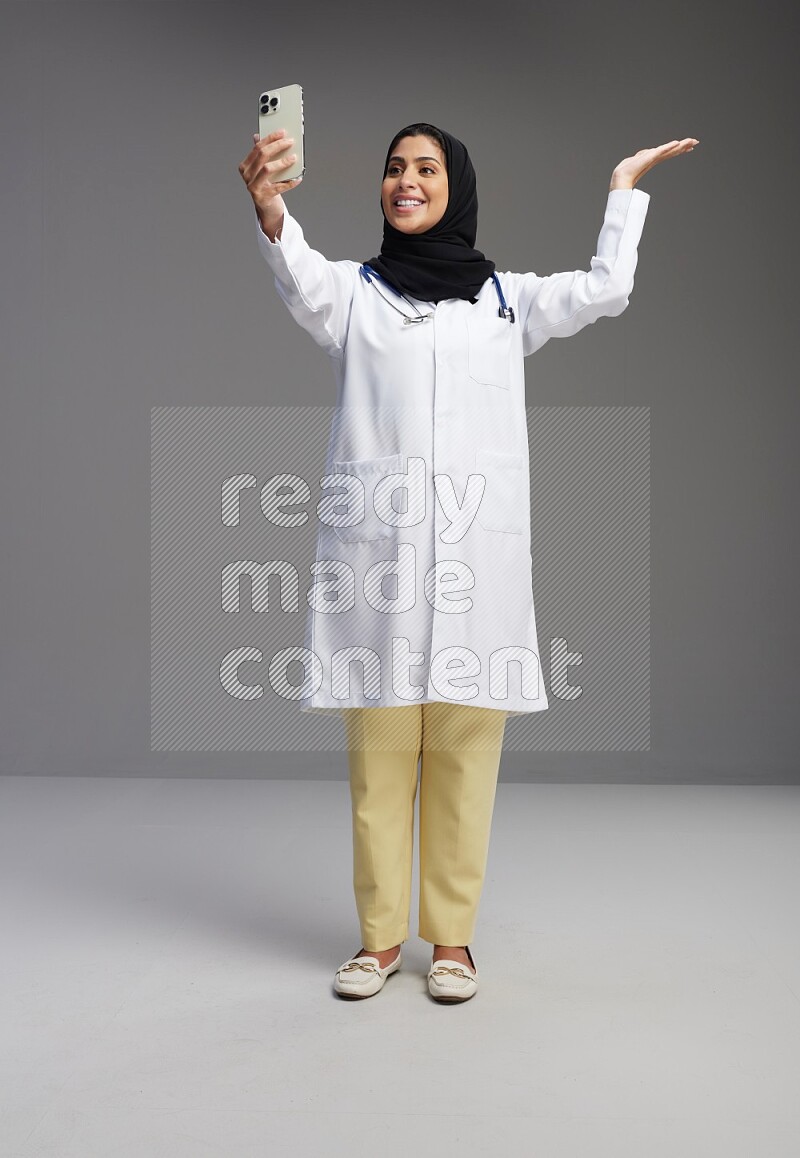 Saudi woman wearing lab coat with stethoscope standing taking selfie on Gray background