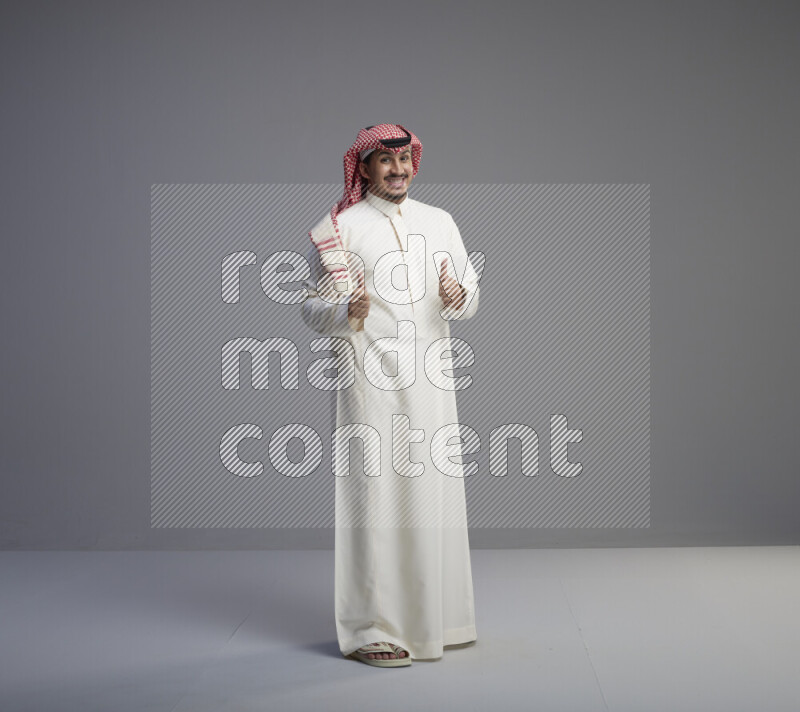 A Saudi man standing wearing thob and red shomag interacting with the camera on gray background