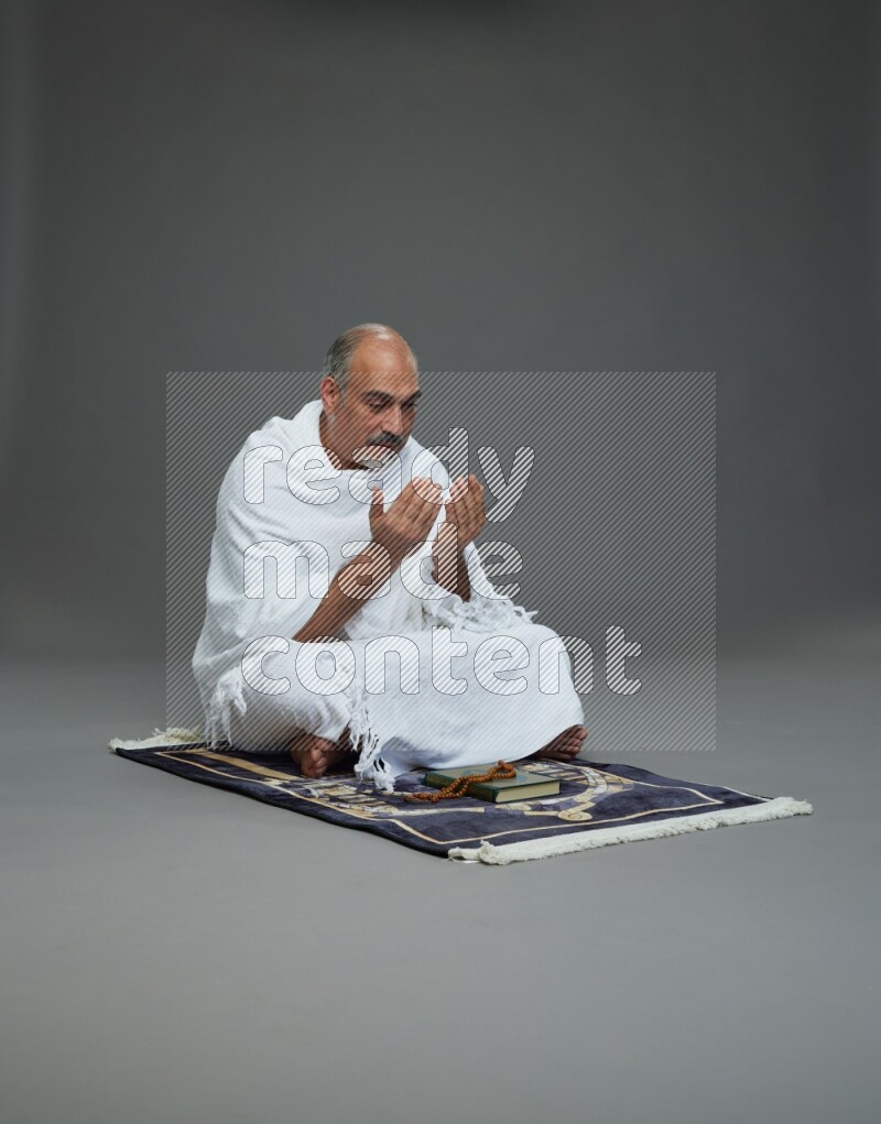 A man wearing Ehram sitting on prayer mat dua'a on gray background