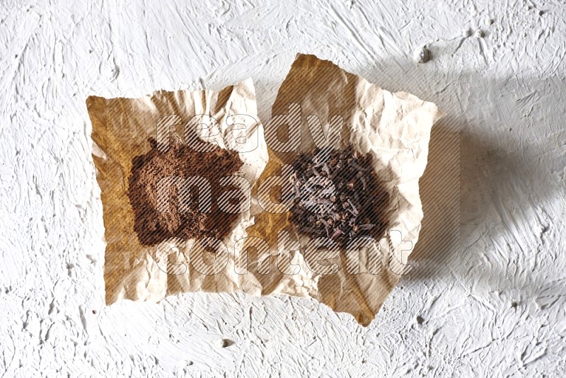 Cloves powder and cloves on 2 crumpled pieces of paper on a textured white flooring