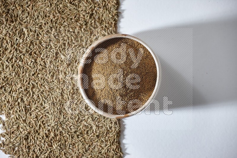 A beige bowl full of cumin powder with cumin seeds beneath it the bowl on a white flooring
