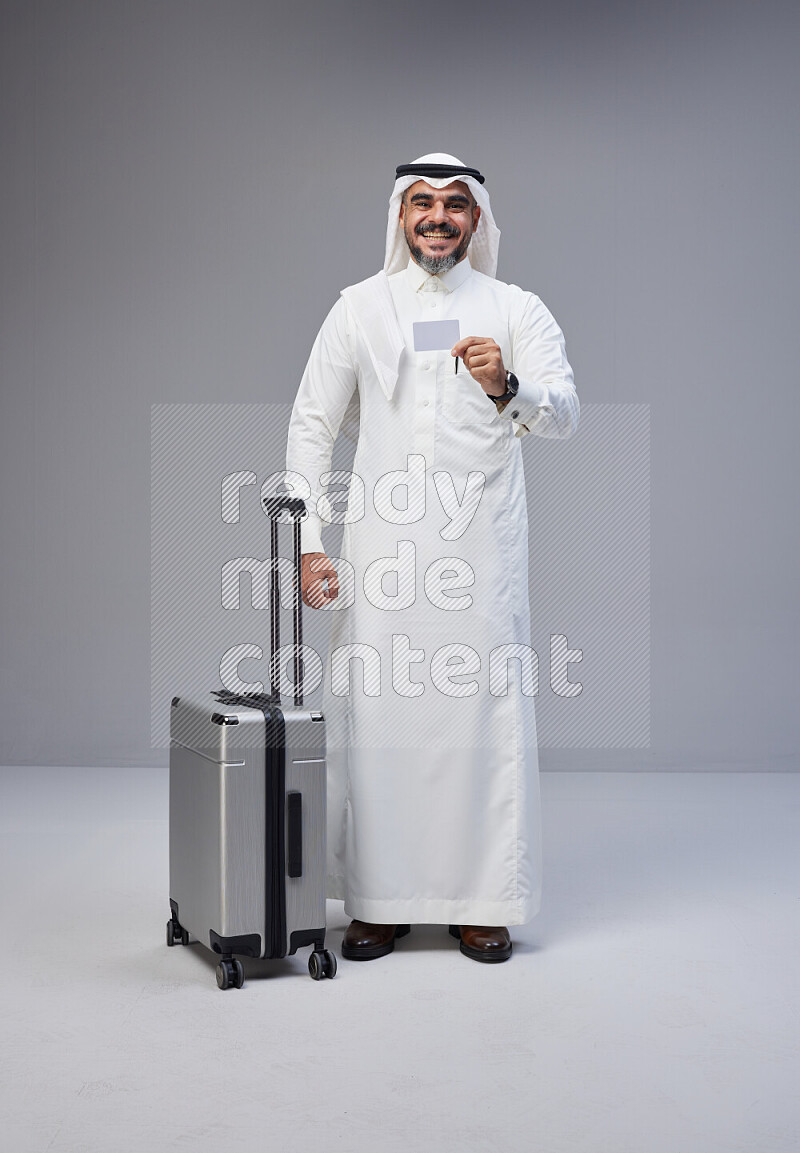 Saudi man wearing Thob and white Shomag standing holding Travel bag and ATM card on Gray background