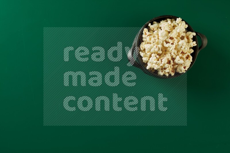 A black ceramic bowl full of popcorn on a green background in a top view shot