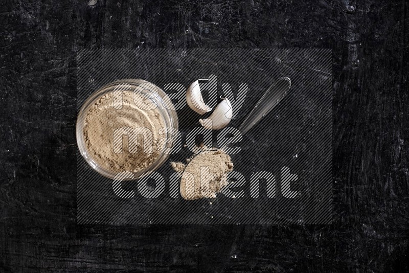 A glass jar full of garlic powder with metal spoon full of the powder on a textured black flooring in different angles