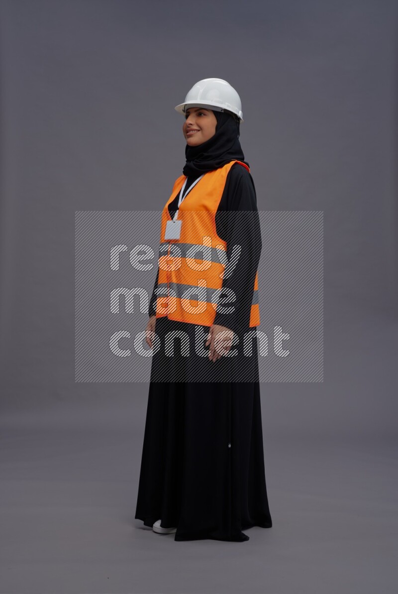 Saudi woman wearing Abaya with engineer vest with neck strap employee badge standing interacting with the camera on gray background