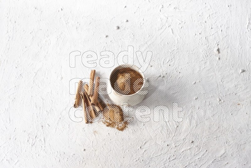Ceramic beige bowl full of cinnamon powder and a metal spoon with cinnamon sticks next of it on a textured white background