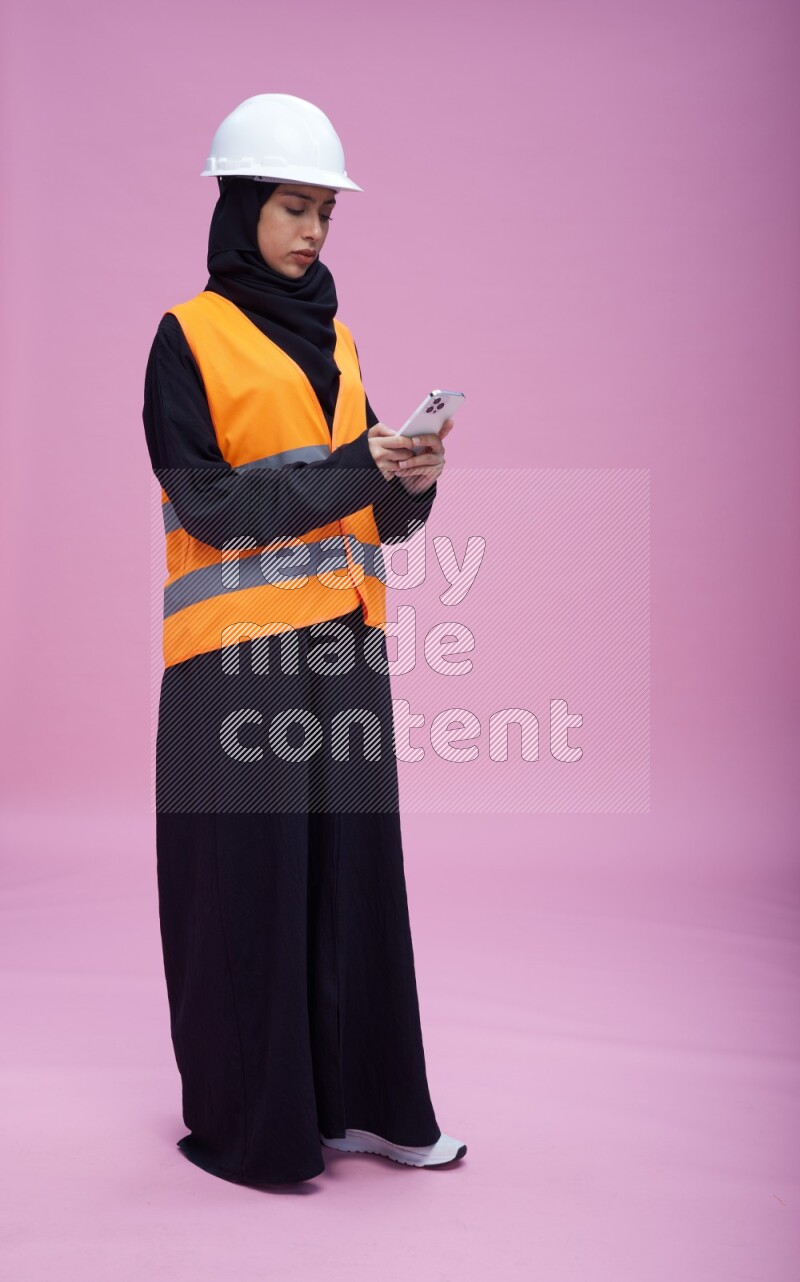 Saudi woman wearing Abaya with engineer vest and helmet standing texting on phone on pink background