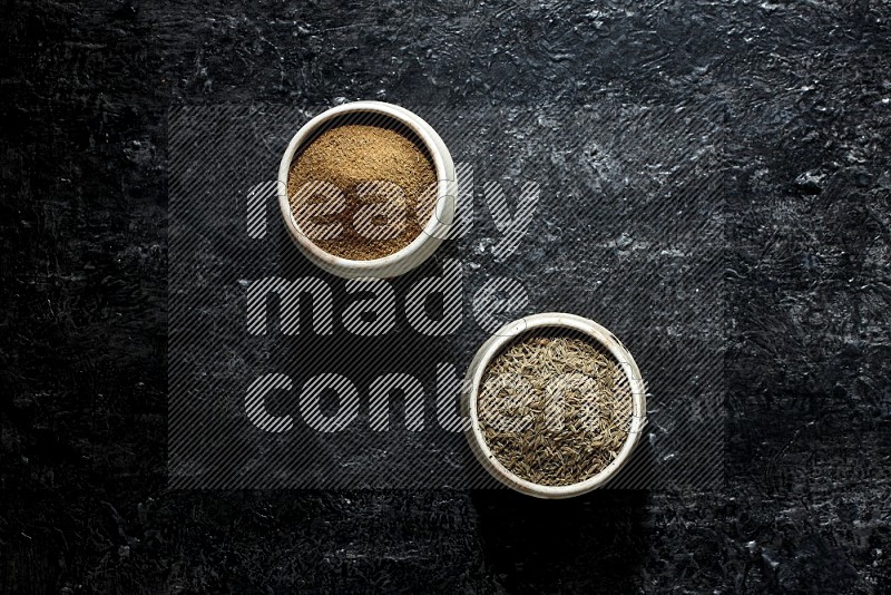 2 beige bowls, one full of cumin powder and the other full of seeds on a textured black flooring