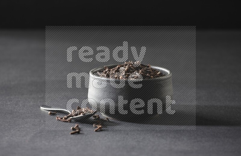 A black pottery bowl full of cloves and a metal spoon next to it on a black flooring